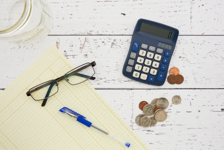 A calculator, coins, eyeglasses and a pen on a desk with a notepad