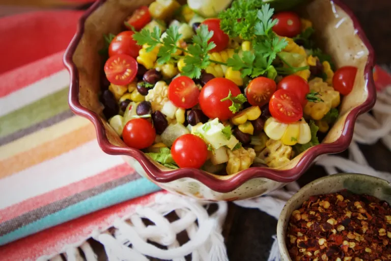 A colorful bowl of salad with tomatoes, corn, beans and parsley on a striped cloth