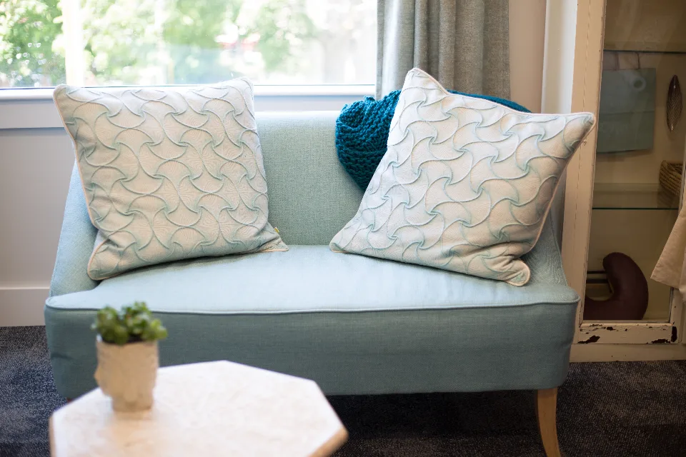 A light blue loveseat with patterned pillows by a sunny window in a cozy living room