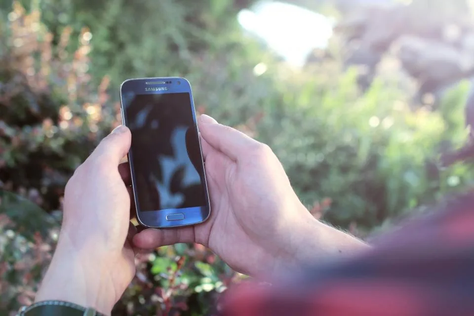 Hands holding an Android phone outdoors near green plants