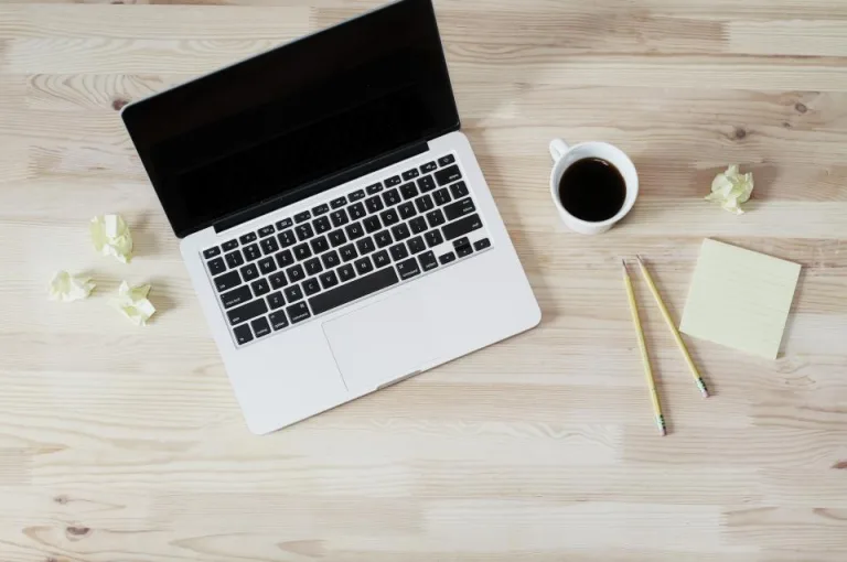 An open laptop, a cup of coffee and a notepad on a light wooden table