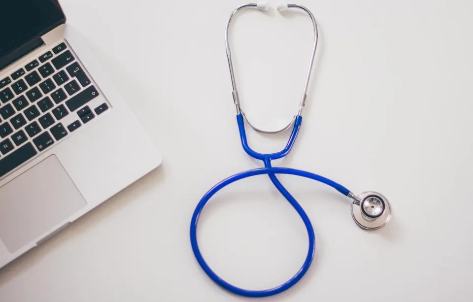 A laptop next to a blue stethoscope on a white desk