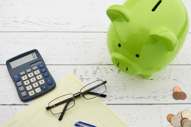 A green piggy bank beside a calculator, eyeglasses and coins