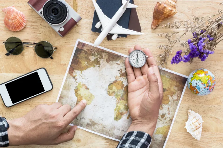 Hands planning a trip over a world map with a compass, sunglasses and a camera