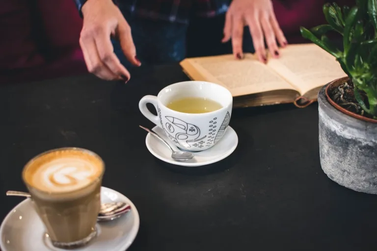 A cup of tea and a coffee on a table beside an open book and a plant
