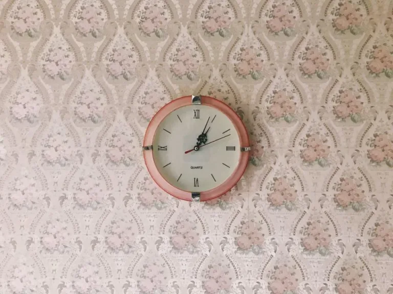 A round wall clock with a pink frame hanging on floral wallpaper