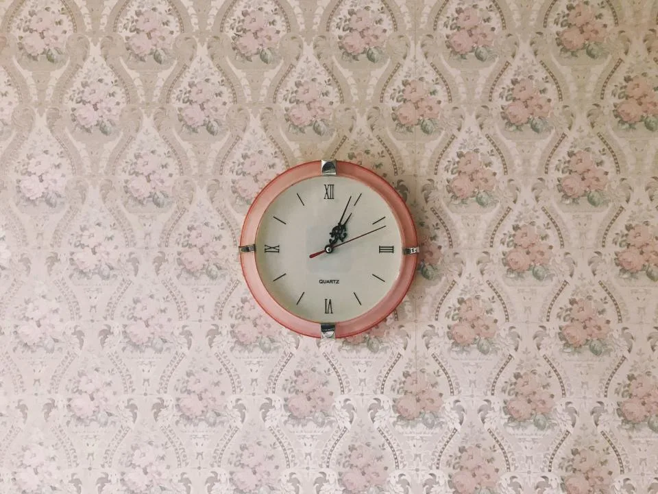 A round wall clock with a pink frame hanging on floral wallpaper