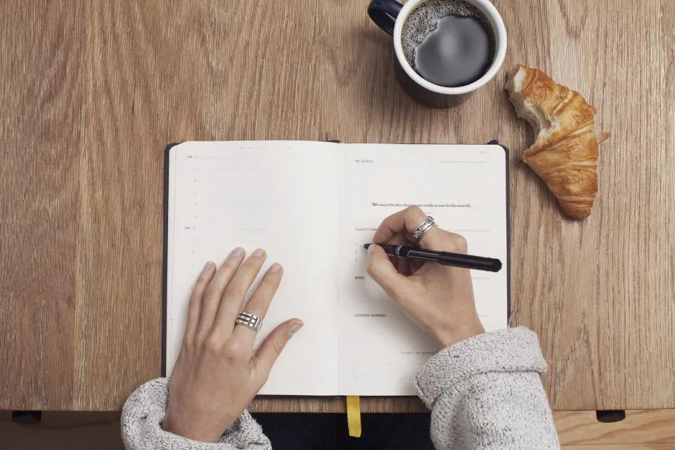 A hand writing in an open notebook beside a cup of coffee and a croissant on a wooden table